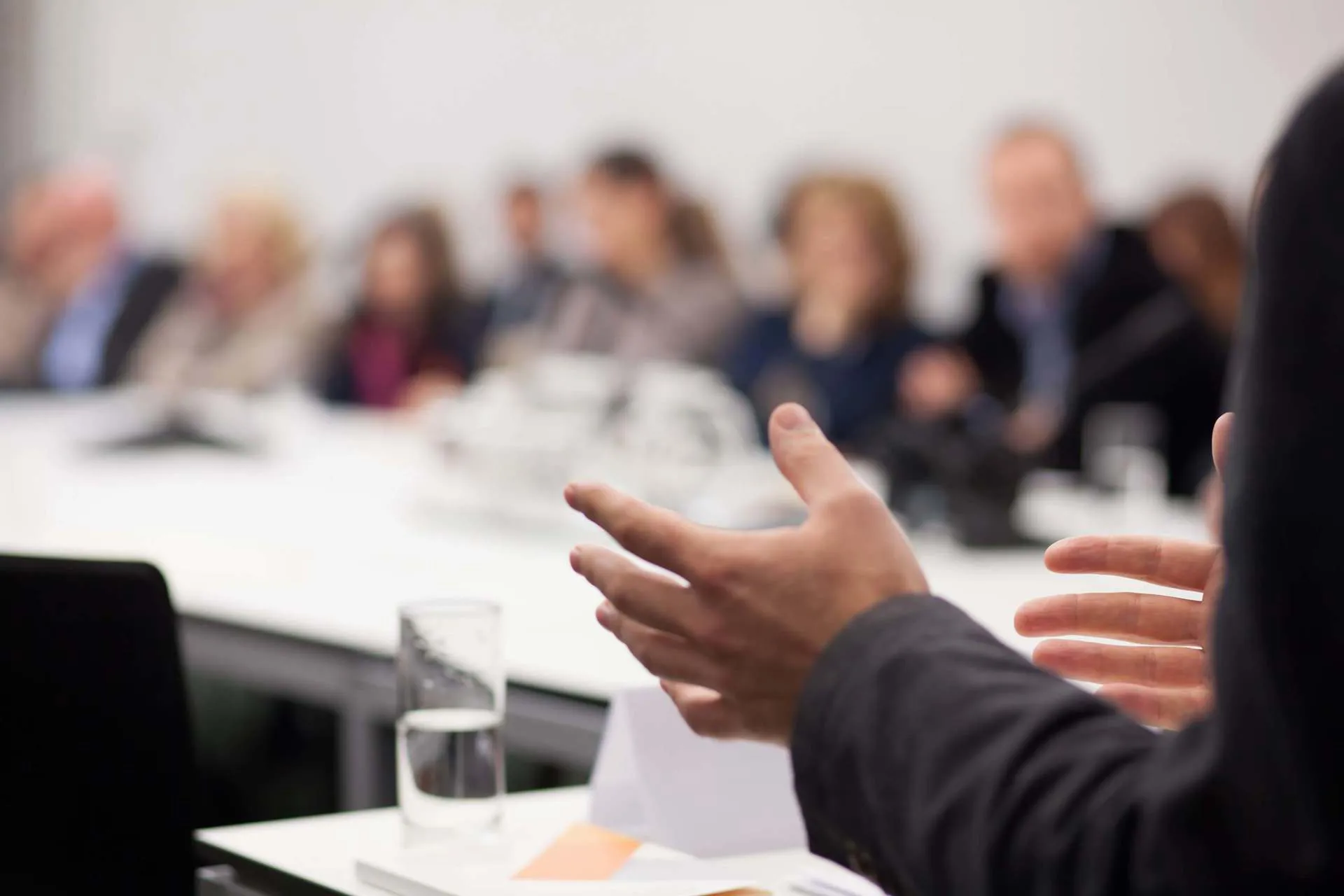 A group of people that have been blurred out look on to someone who is using hand gestures to explain something at a business meeting.