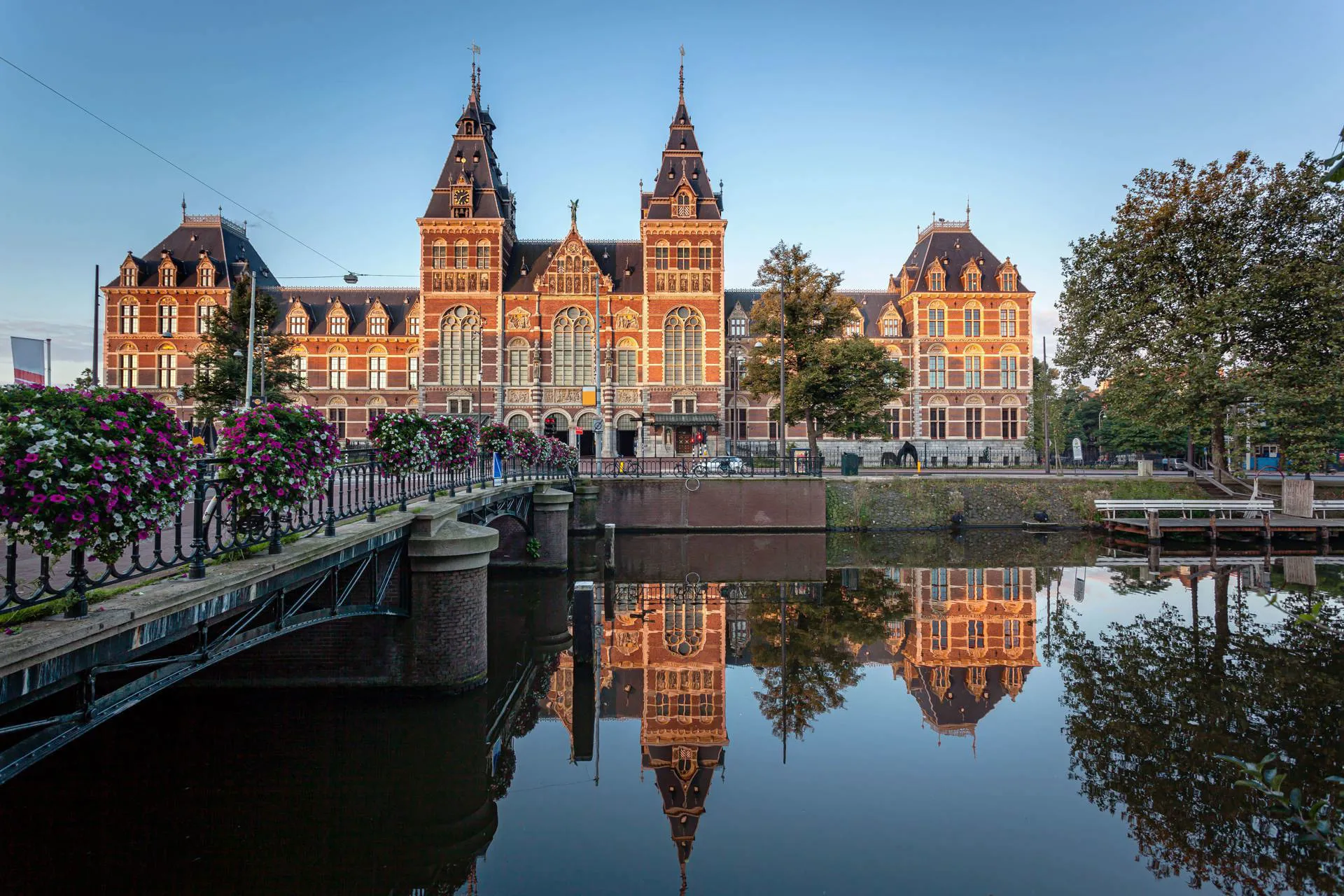 A sunlit Rijksmuseum in Amsterdam as a bridge leads up to it with pink and white flowers.