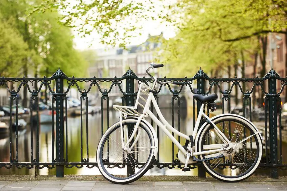 A white bicycle leant against a metal railing that is overlooking a river in Amsterdam.
