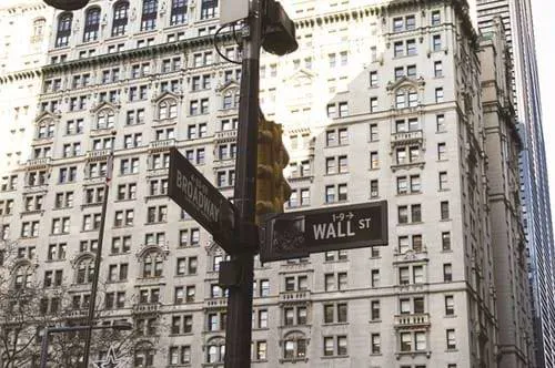 New York City sign reading Wall St and Broadway, with a tall stone building behind.