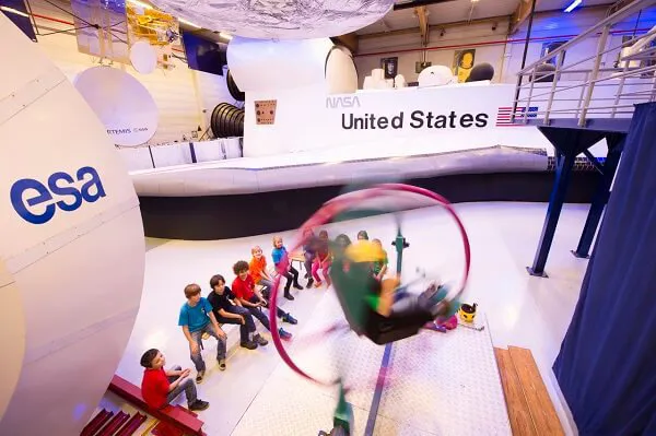 A group of students in multicoloured shirts look on as a student takes part in a physical activity at the Euro Space Center.
