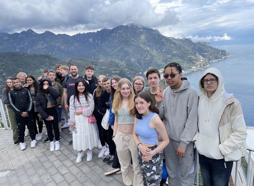 Students on a school trip to Italy, with the sea and mountains behind the group.