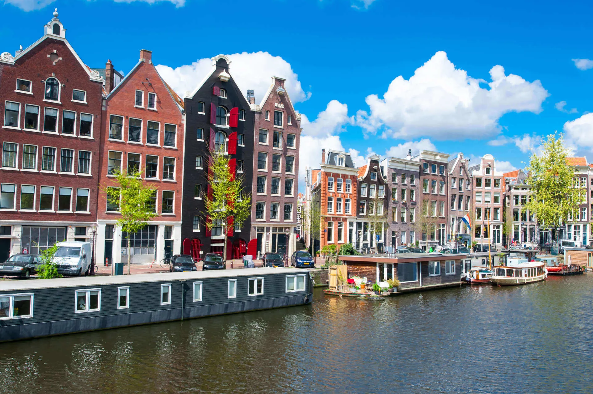 Tall pointed buildings side a river in Amsterdam as green trees and canal boats line the riverbank.