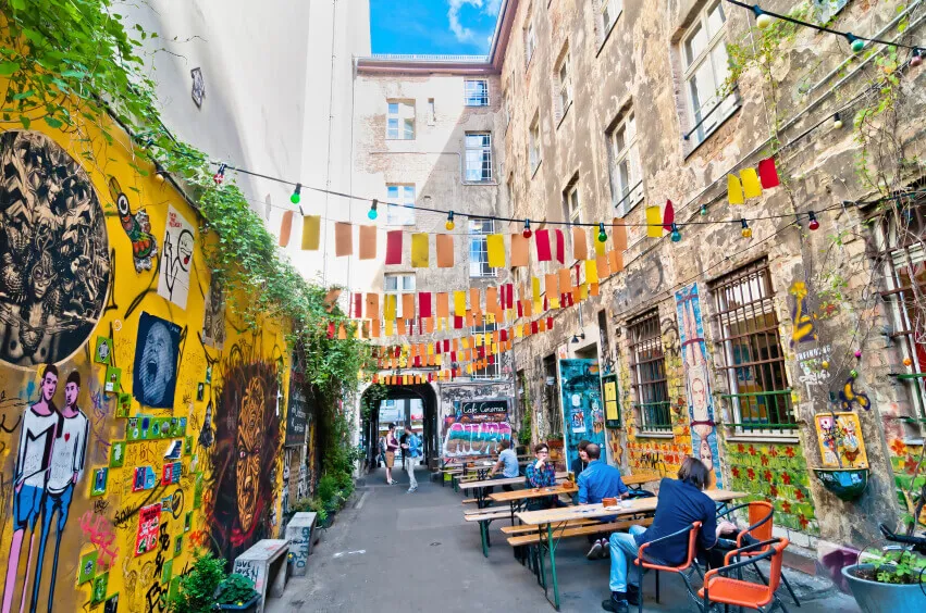 A side street in the city of Berlin, decorated with coloured flags, seating, plants and street art up the left wall.